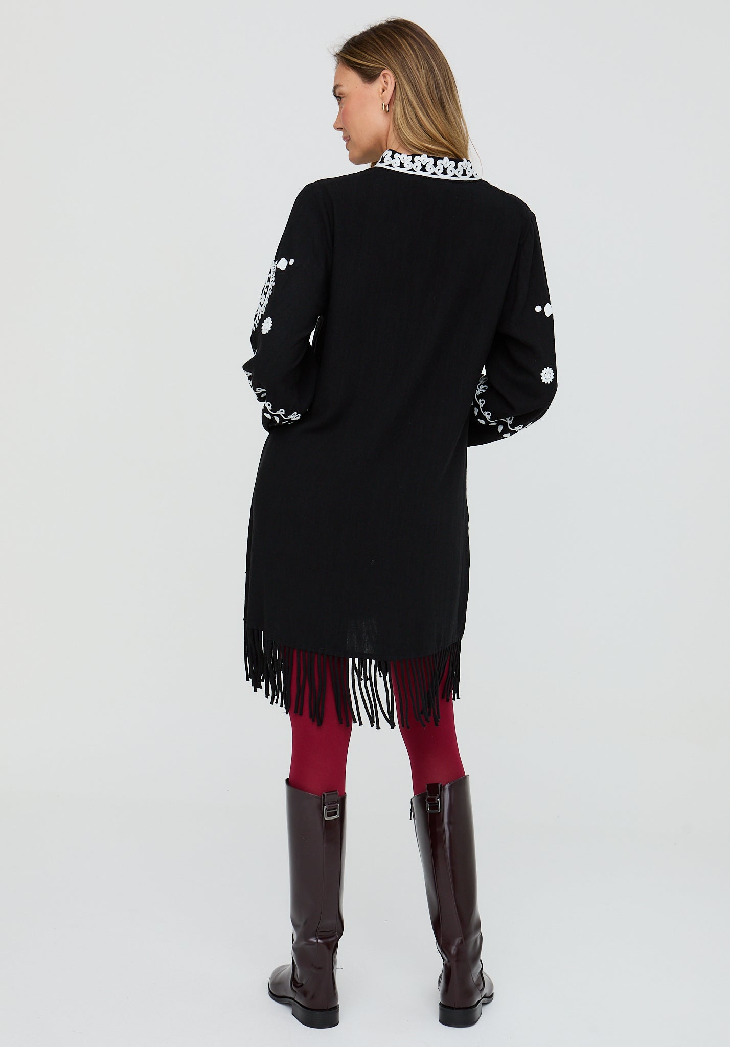 Woman wearing a black cardigan with white patterns and fringes, red pants, and dark boots on a light gray background.