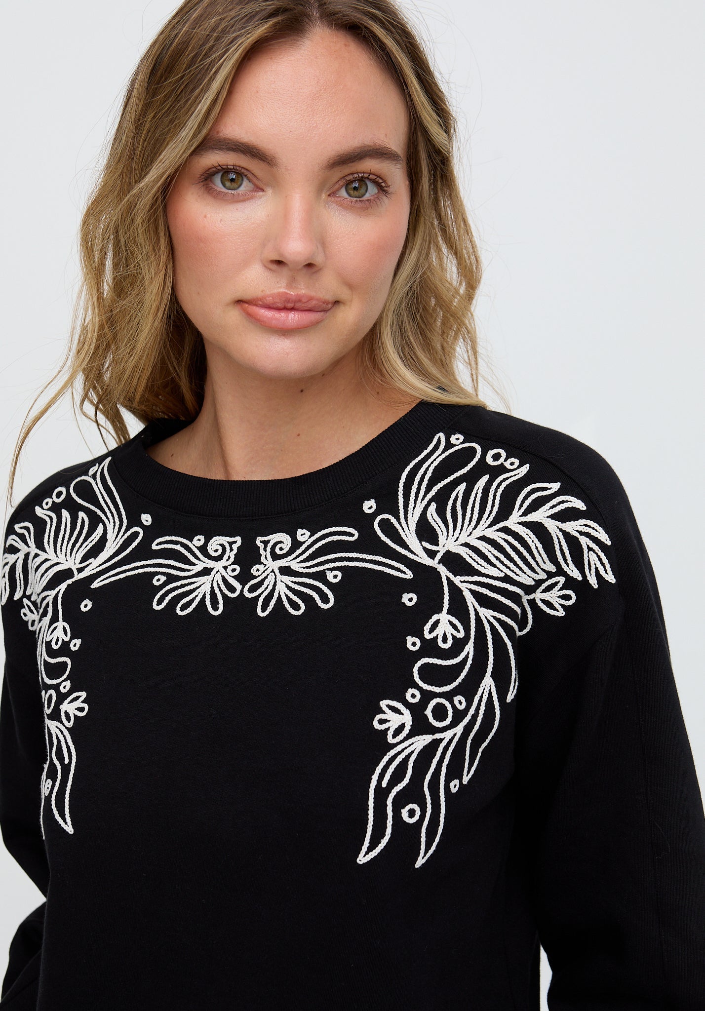 Woman wearing a black sweater with white floral embroidery on a plain background