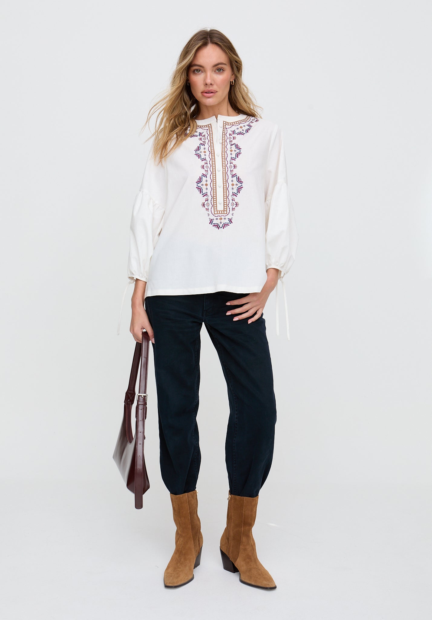 Woman wearing a white embroidered blouse with black pants and brown boots on a white background