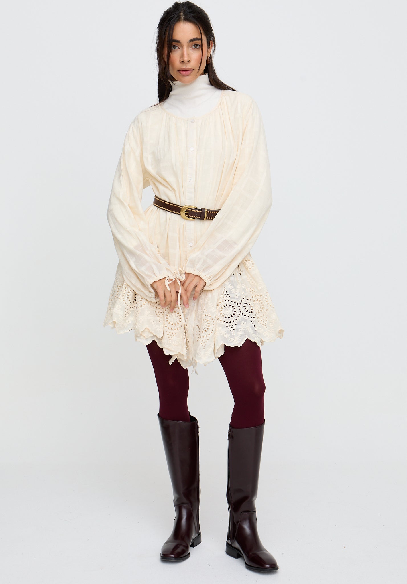 Woman wearing a cream lace dress with burgundy leggings and boots on a white background