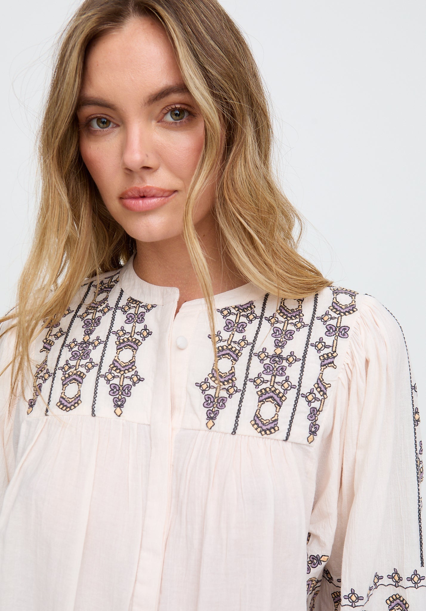 Woman wearing a white blouse with intricate black and purple embroidery on a plain background