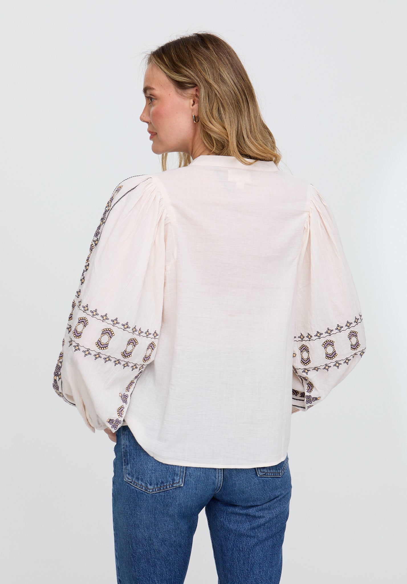 Woman wearing a white blouse with embroidered sleeves and blue jeans on a light gray background