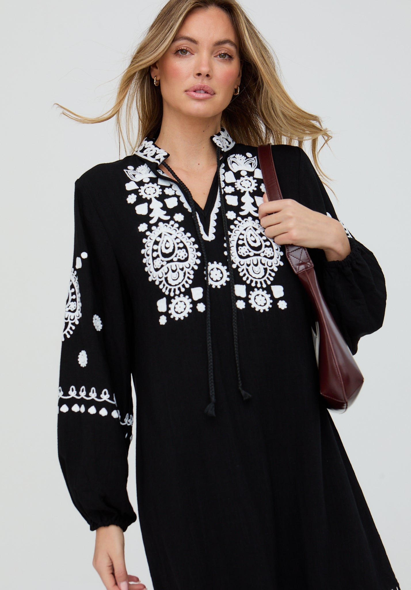 Woman wearing a black dress with white embroidery holding a brown bag against a light gray background