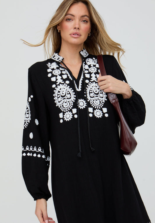 Woman wearing a black dress with white embroidery holding a brown bag against a light gray background