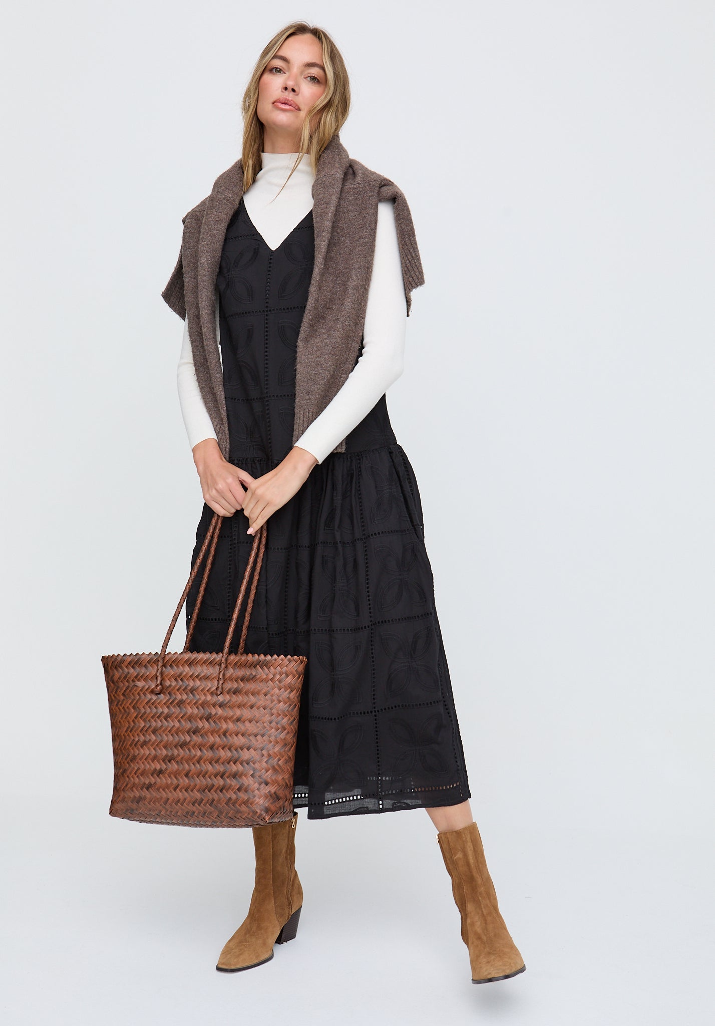 Woman wearing a black dress, white sweater, brown vest, and holding a woven bag on a white background