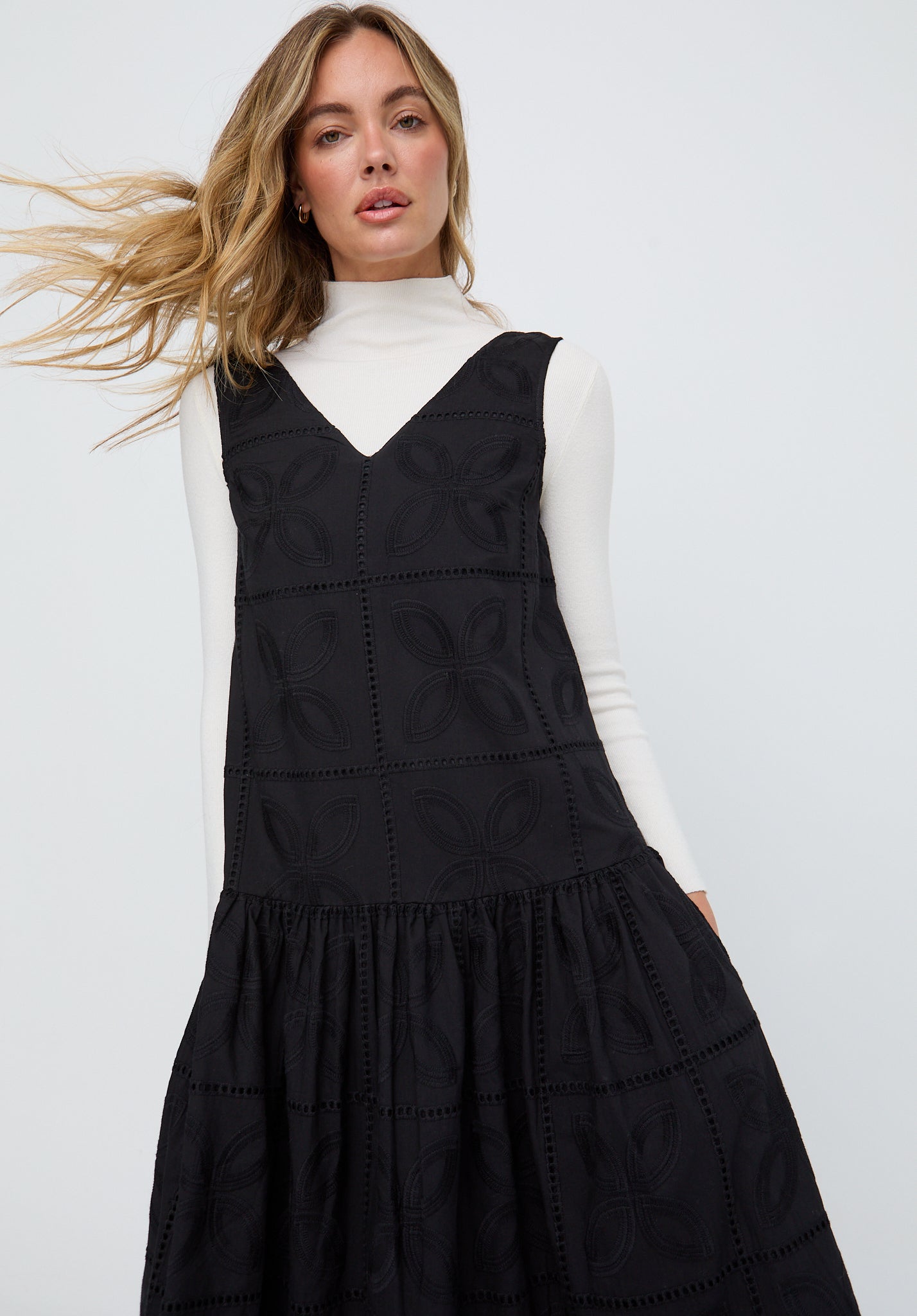 Woman wearing a black pinafore dress over a white turtleneck on a light gray background