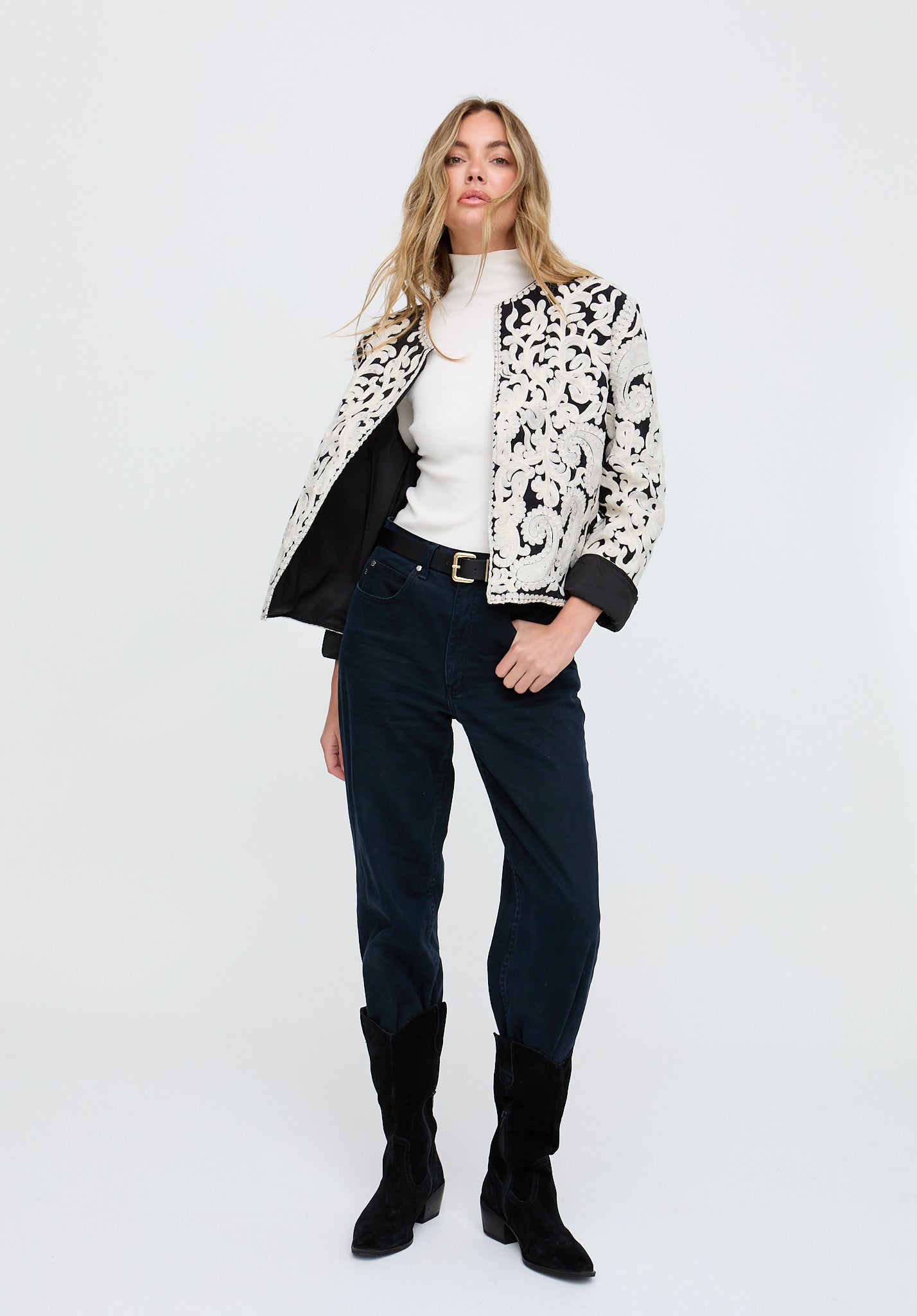 Woman wearing a patterned coat, white shirt, black pants, and black boots on a white background