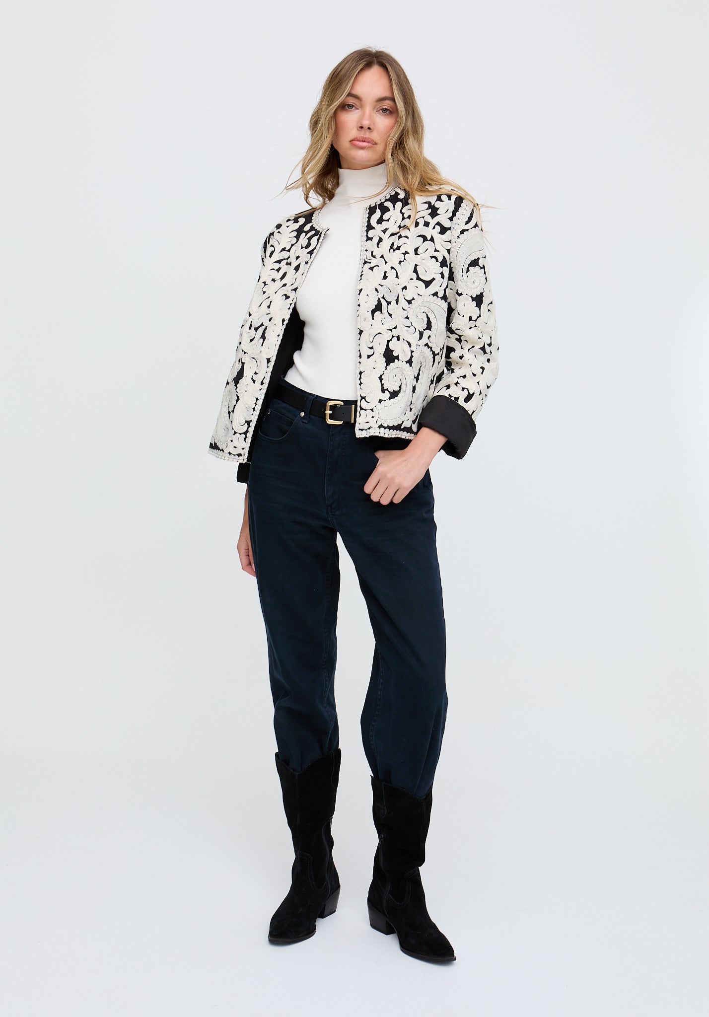 Woman wearing a patterned jacket, white turtleneck, dark jeans, and black boots on a white background