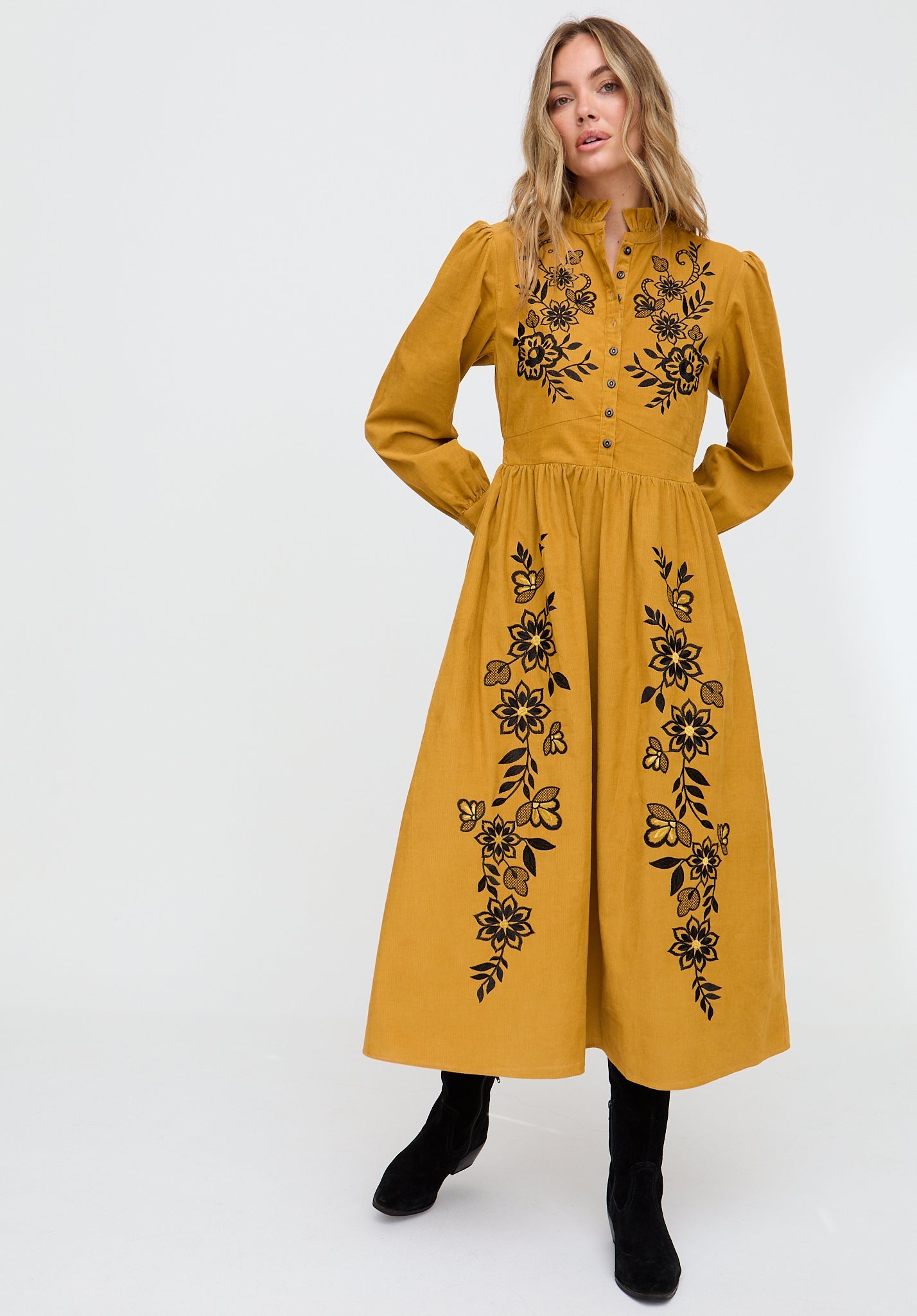 Woman wearing a mustard yellow dress with black floral embroidery on a white background