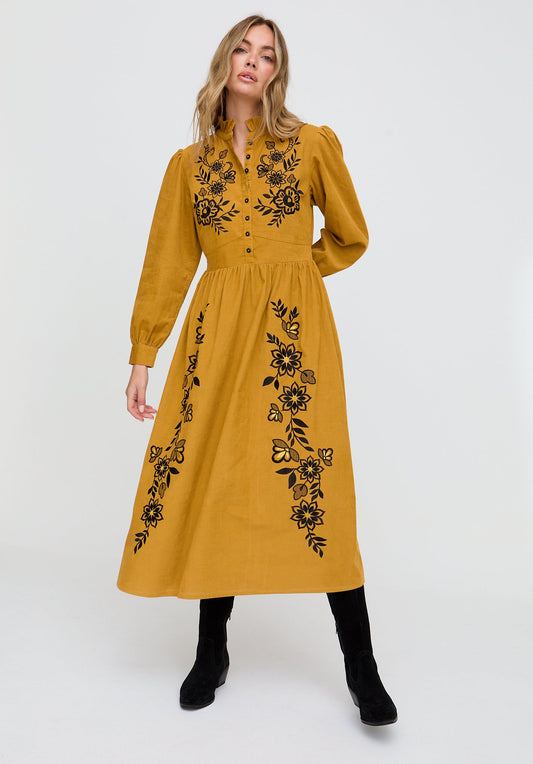 Woman wearing a mustard yellow dress with black floral embroidery on a white background