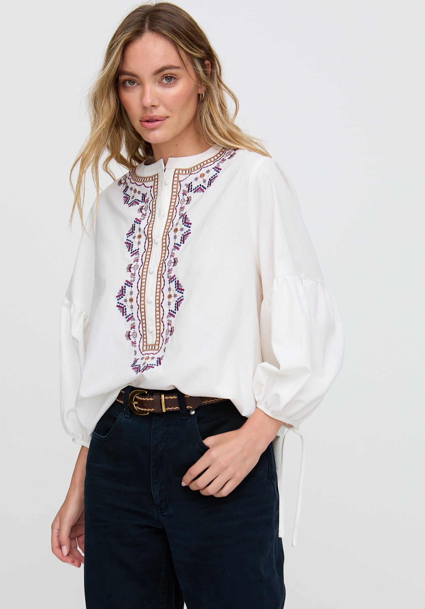 Woman wearing a white blouse with floral embroidery and dark pants on a plain background