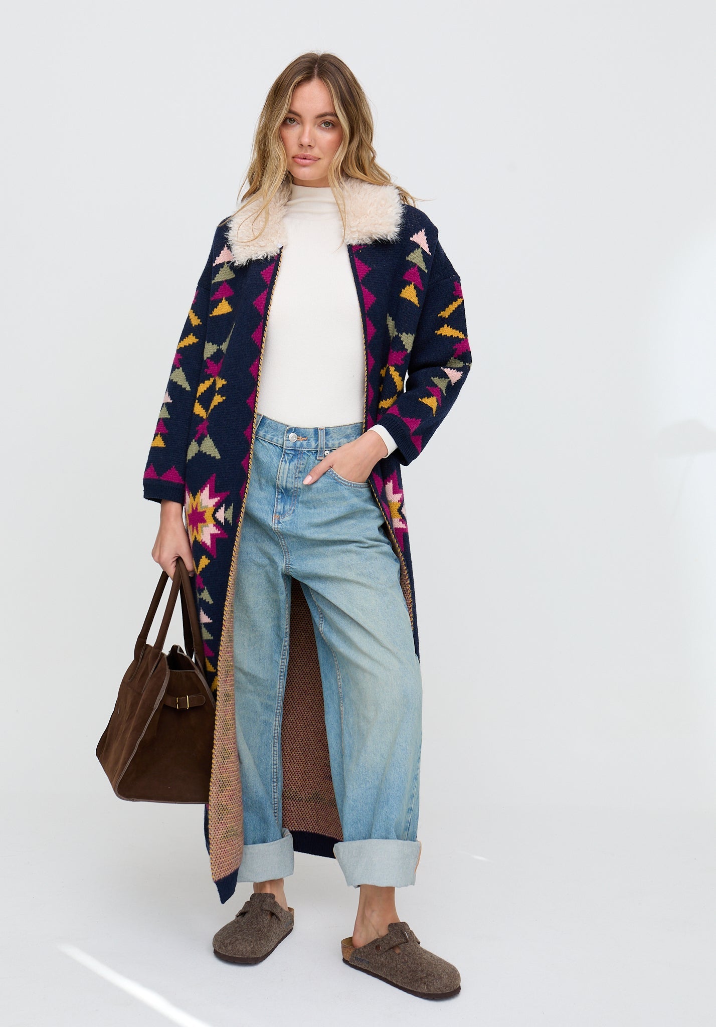 Woman wearing a colorful coat with a pattern, holding a brown bag on a white background