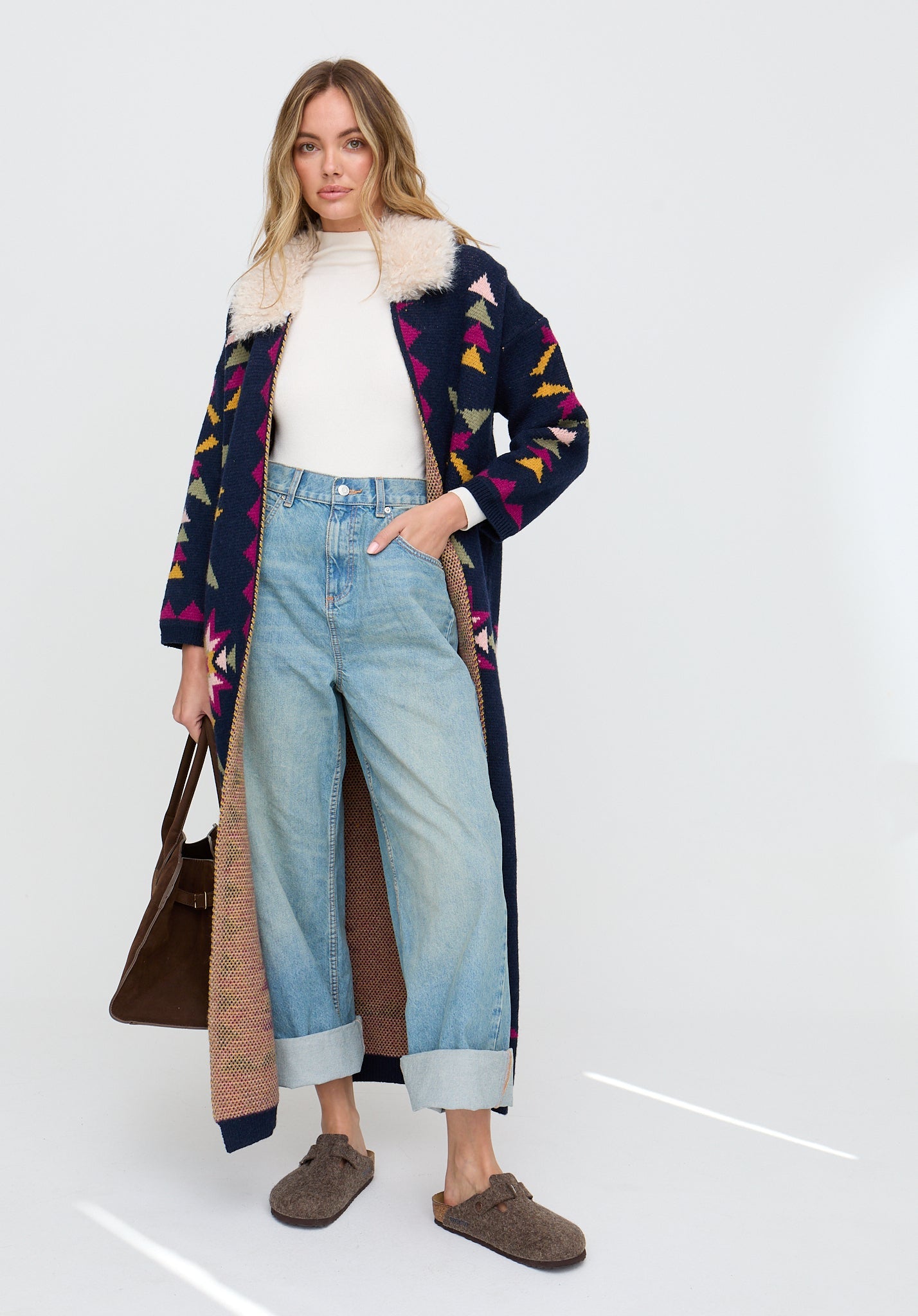 Woman wearing a patterned coat with a fur-lined hood, white sweater, blue jeans, and brown shoes on a white background.