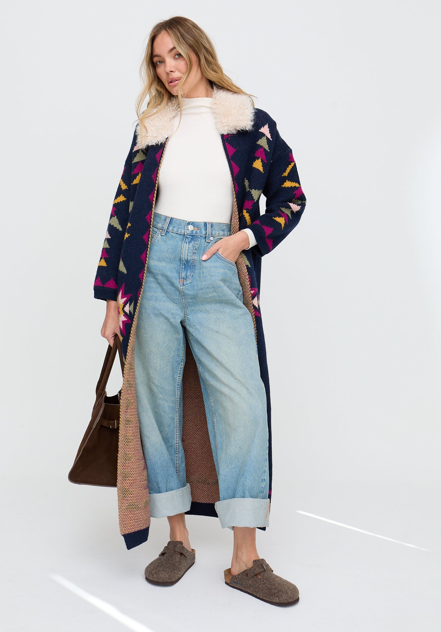 Woman wearing a patterned coat, white sweater, blue jeans, and brown shoes on a white background