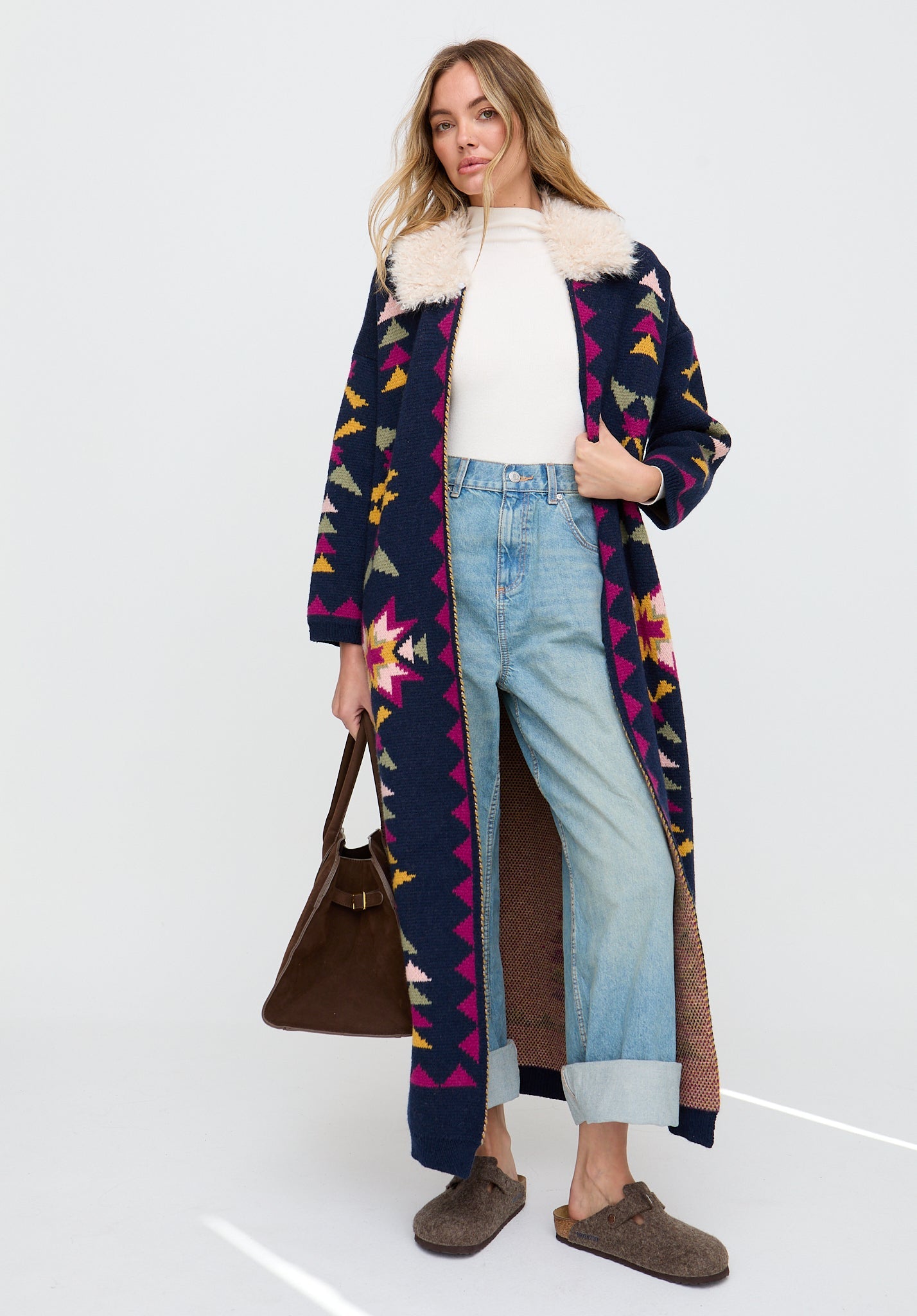 Woman wearing a long, patterned coat over a white sweater and jeans, holding a brown bag on a white background.