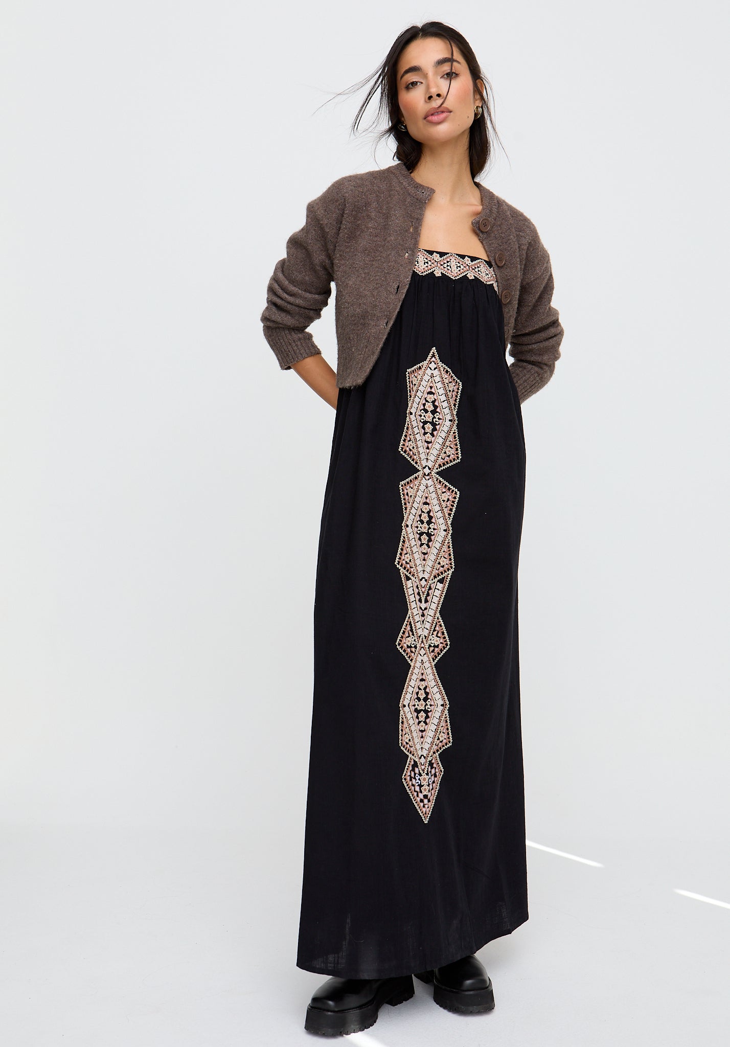 Woman wearing a black dress with intricate gold embroidery and a brown cardigan on a white background