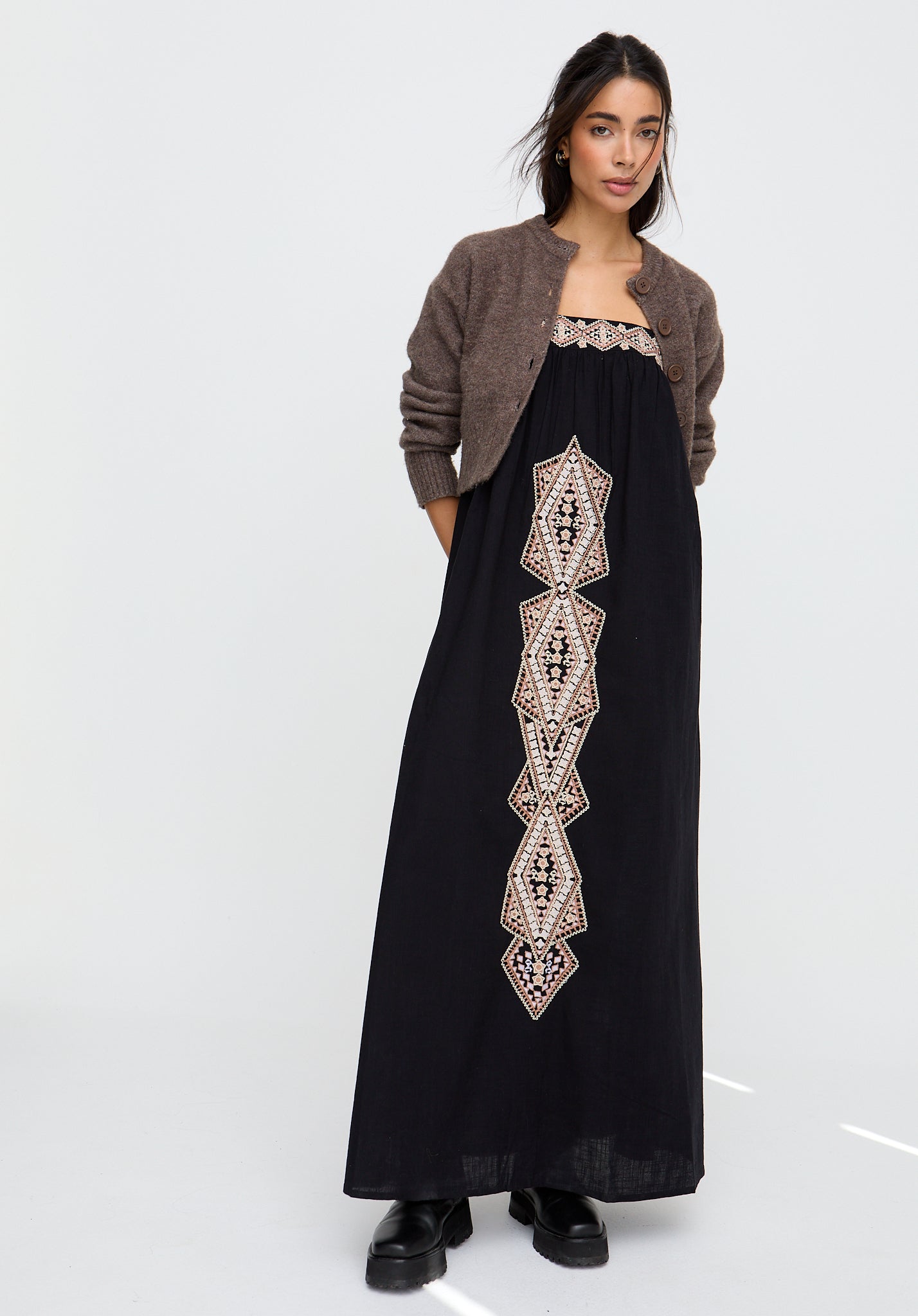 Woman wearing a black dress with geometric embroidery and a brown cardigan on a white background