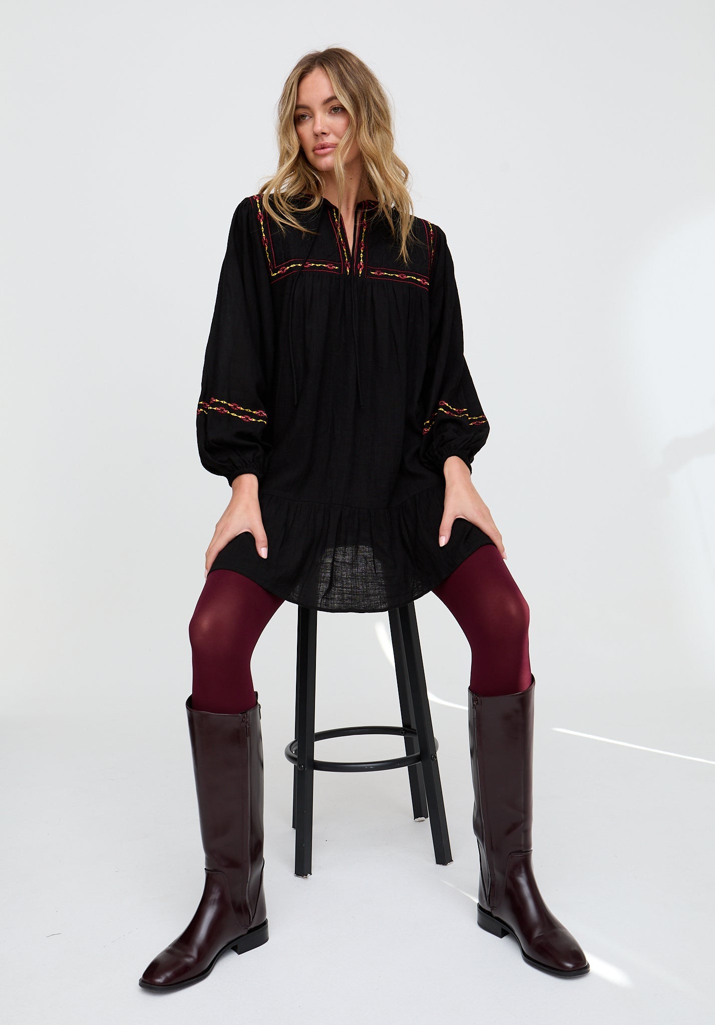 Woman sitting on a stool wearing a black dress with red embroidery, maroon tights, and brown knee-high boots.