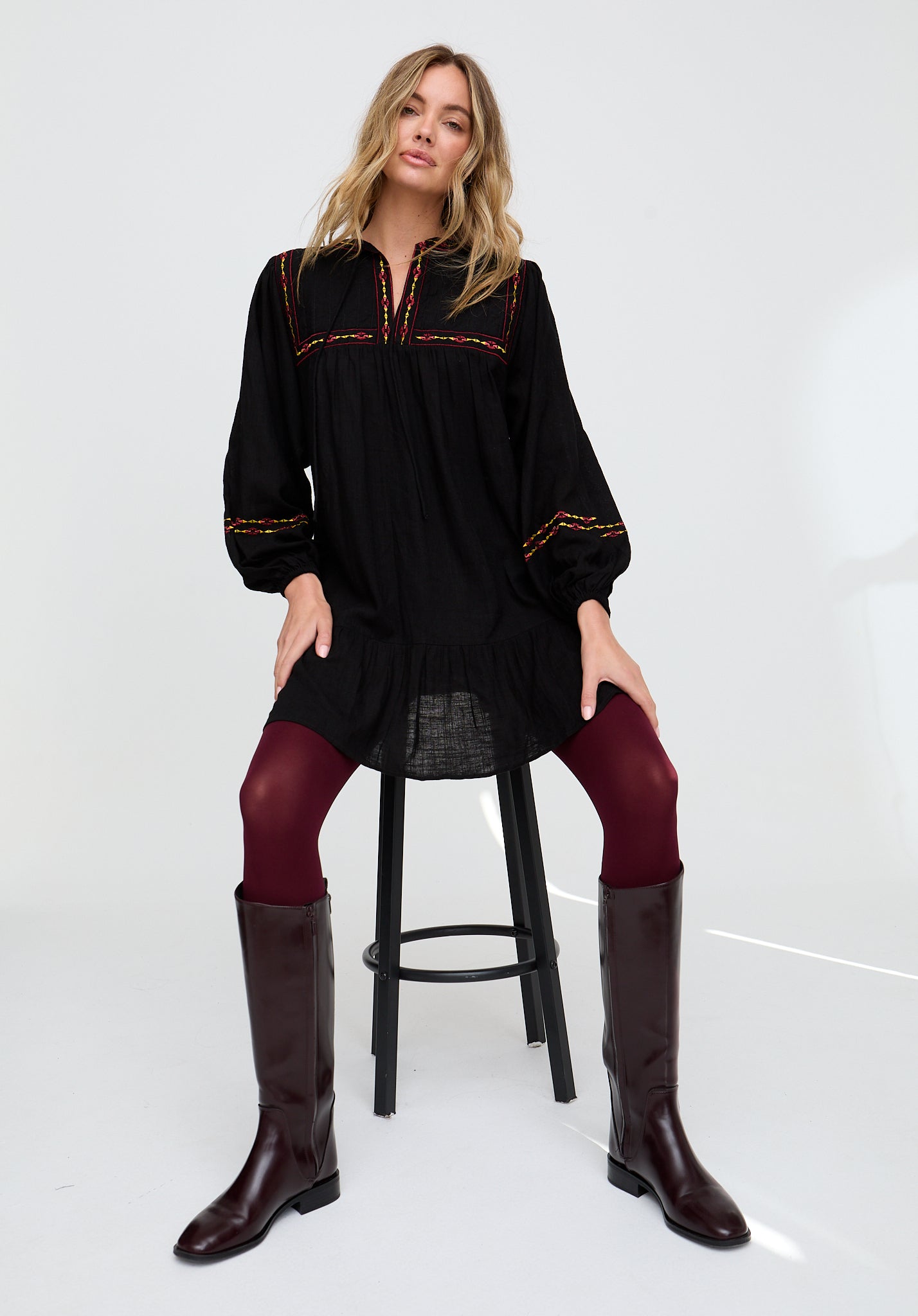 Woman wearing a black long-sleeve top with red embroidery, black leggings, and brown knee-high boots on a white background.