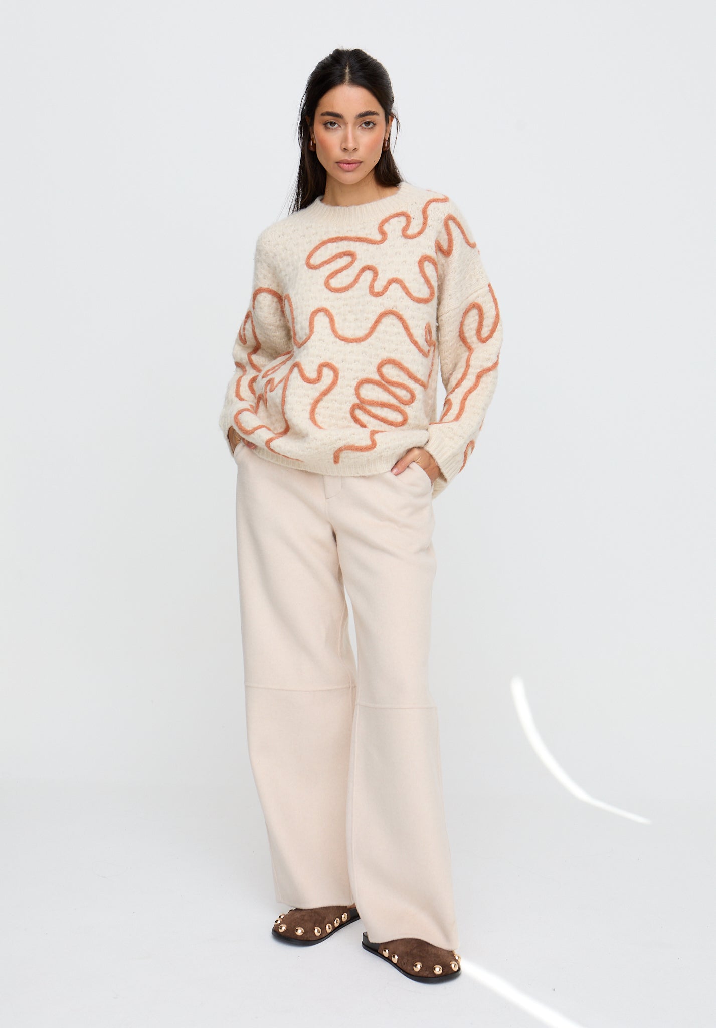 Woman wearing a cream sweater with orange abstract patterns and beige pants on a white background