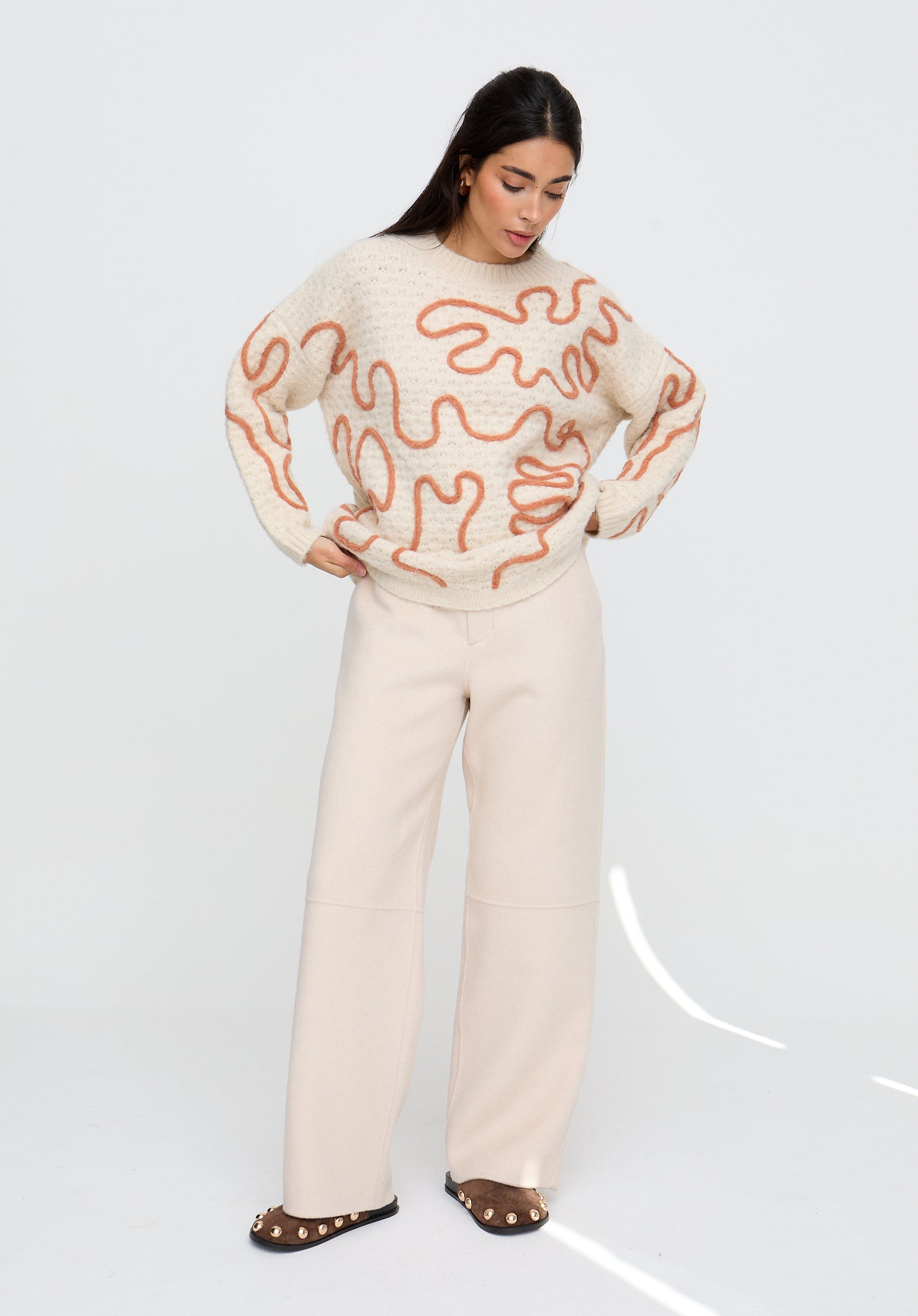 Woman wearing a cream sweater with red abstract patterns and white pants on a light gray background