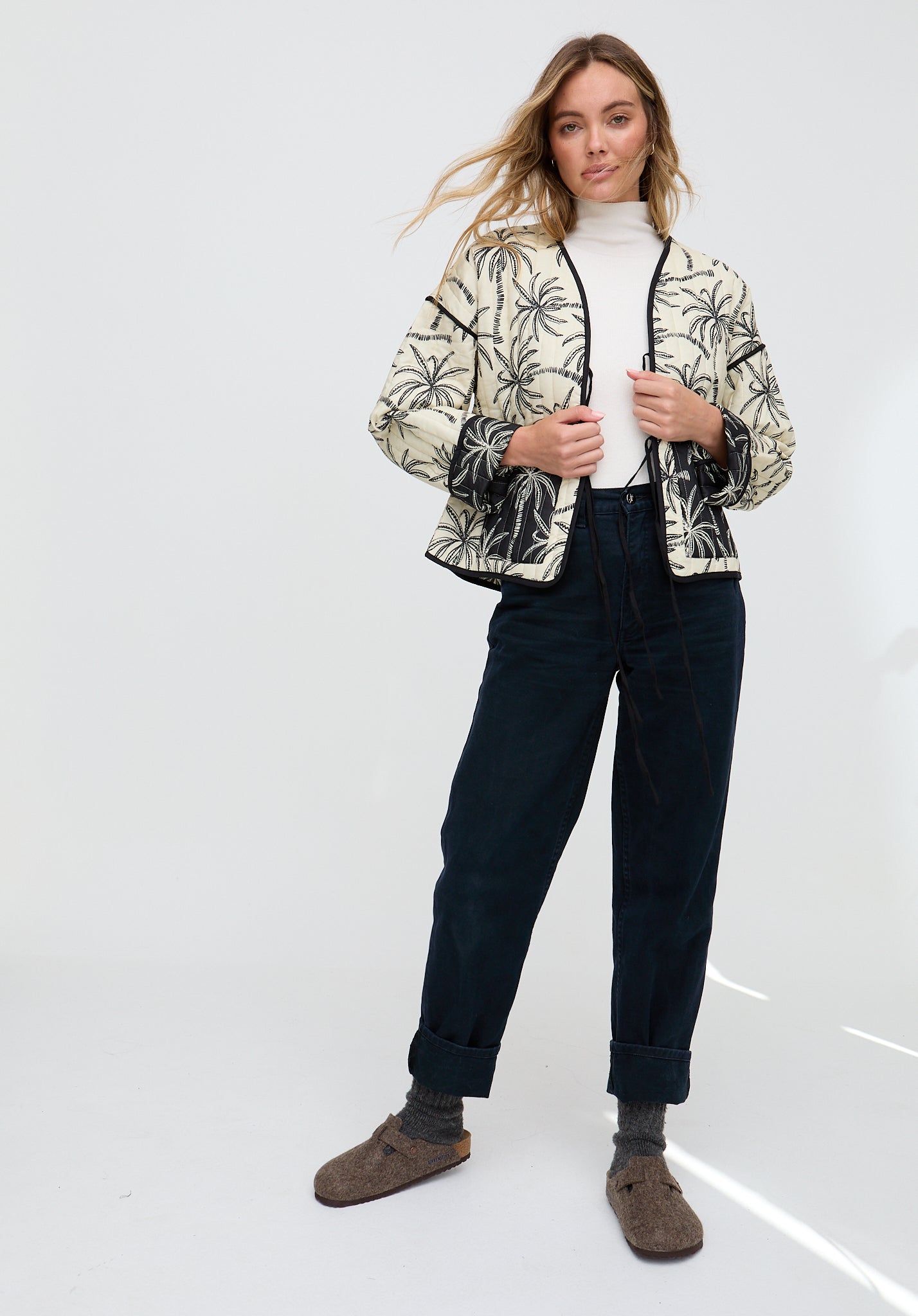 Woman wearing a patterned jacket, white turtleneck, dark jeans, and brown boots on a white background