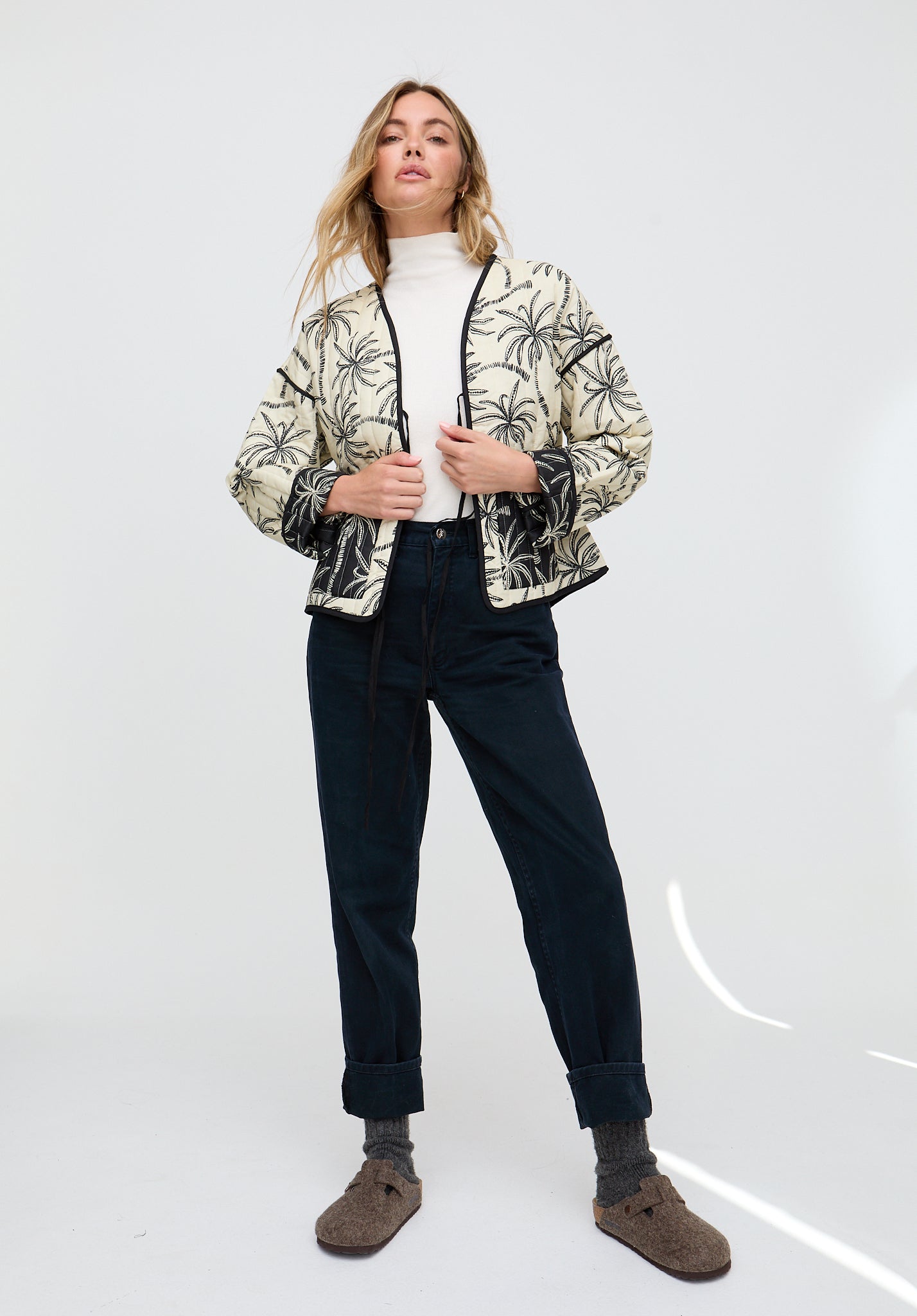 Woman wearing a patterned jacket, white turtleneck, dark pants, and brown shoes on a white background