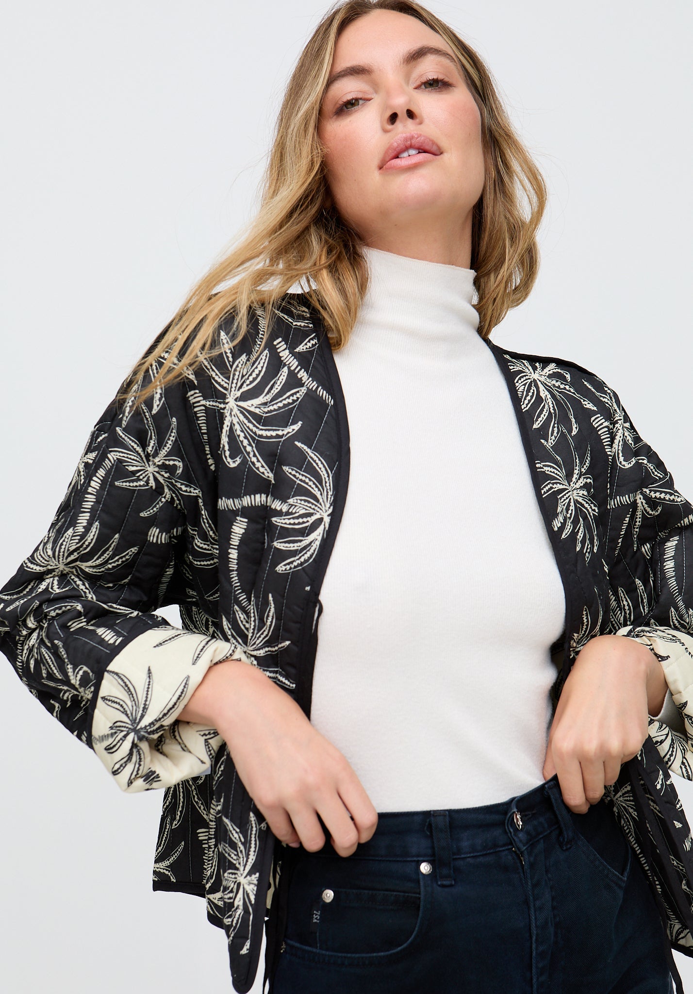 Woman wearing a black floral patterned cardigan over a white turtleneck and dark jeans on a white background
