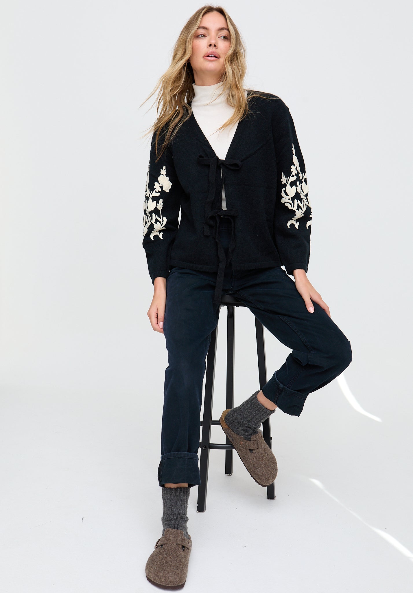 Woman wearing a black cardigan with white floral embroidery, sitting on a stool against a white background.