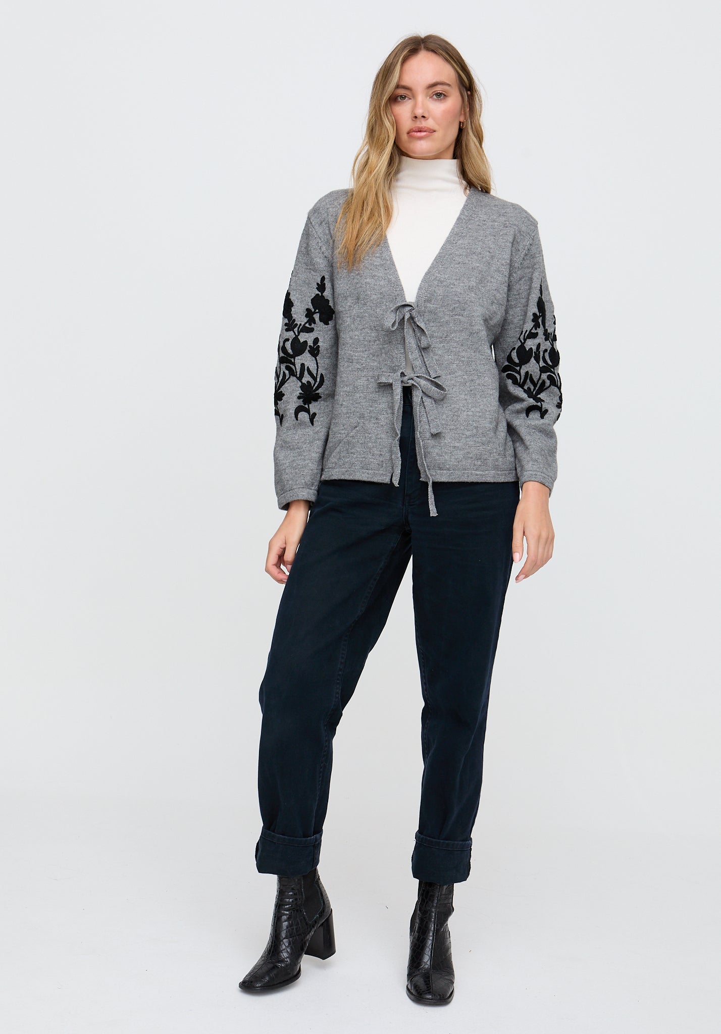 Woman wearing a gray cardigan with black floral patterns, white turtleneck, and black pants on a white background