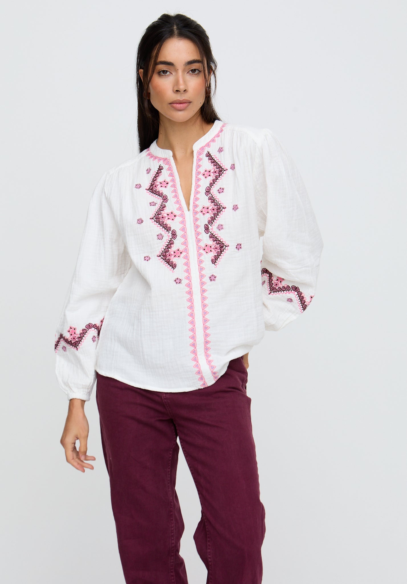 Woman wearing a white blouse with pink embroidery and maroon pants on a light gray background