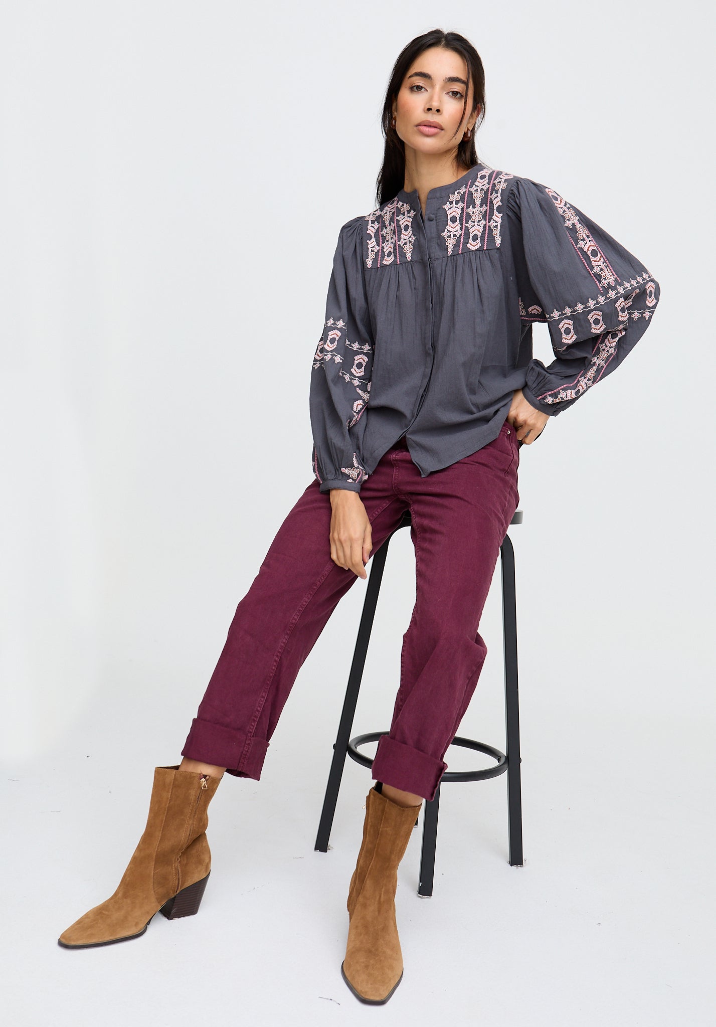 Woman sitting on a stool wearing a patterned blouse and maroon pants with brown boots on a white background