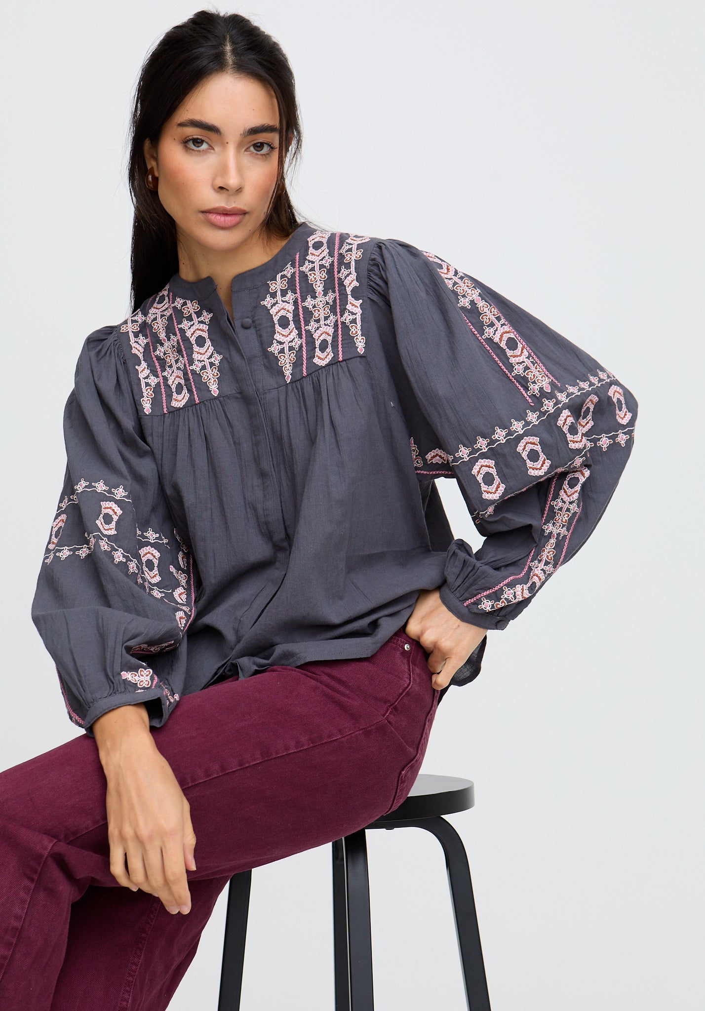 Woman wearing a dark gray blouse with intricate pink embroidery sitting on a stool against a white background