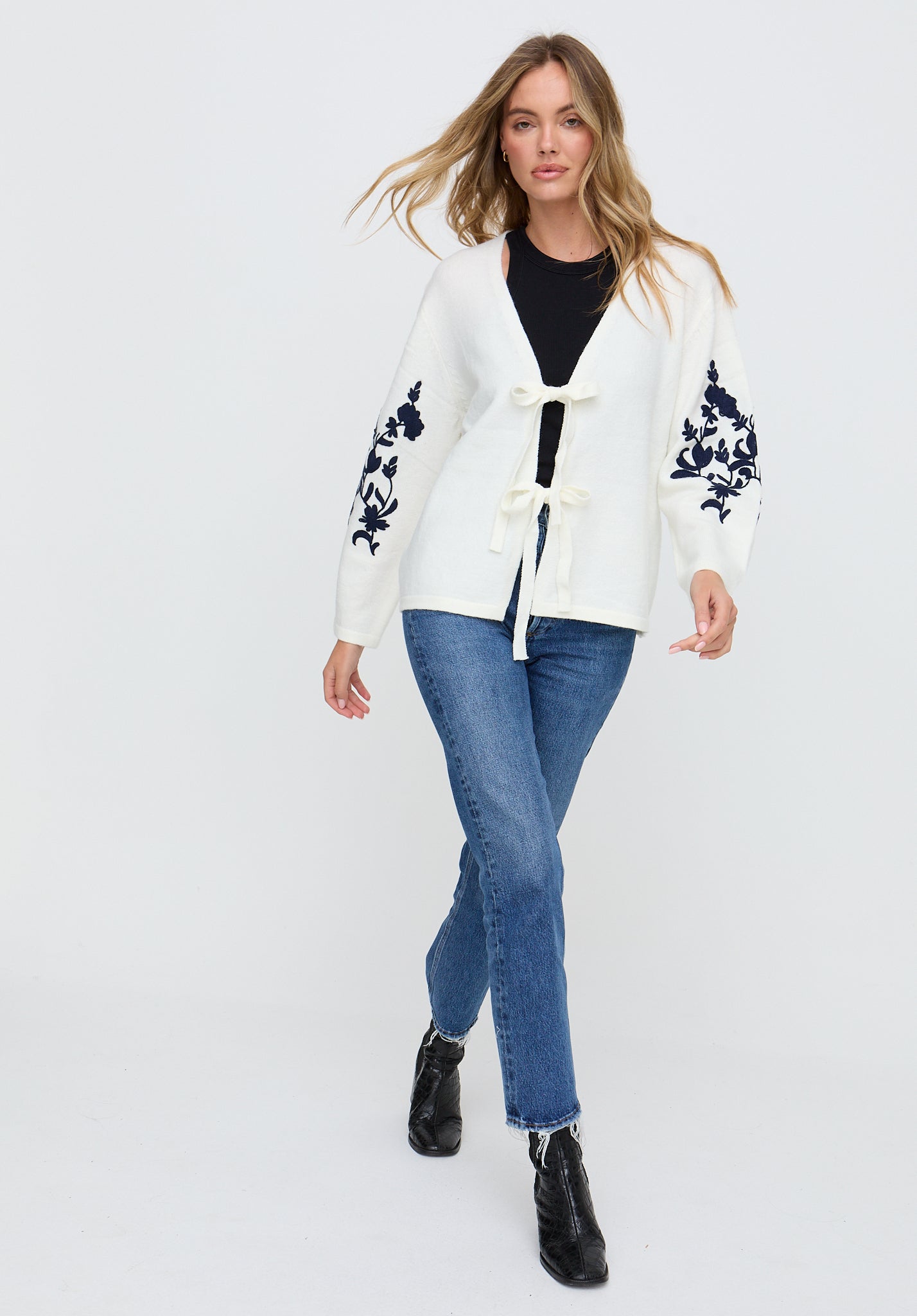 Woman wearing a white cardigan with floral patterns, black top, blue jeans, and black boots on a white background
