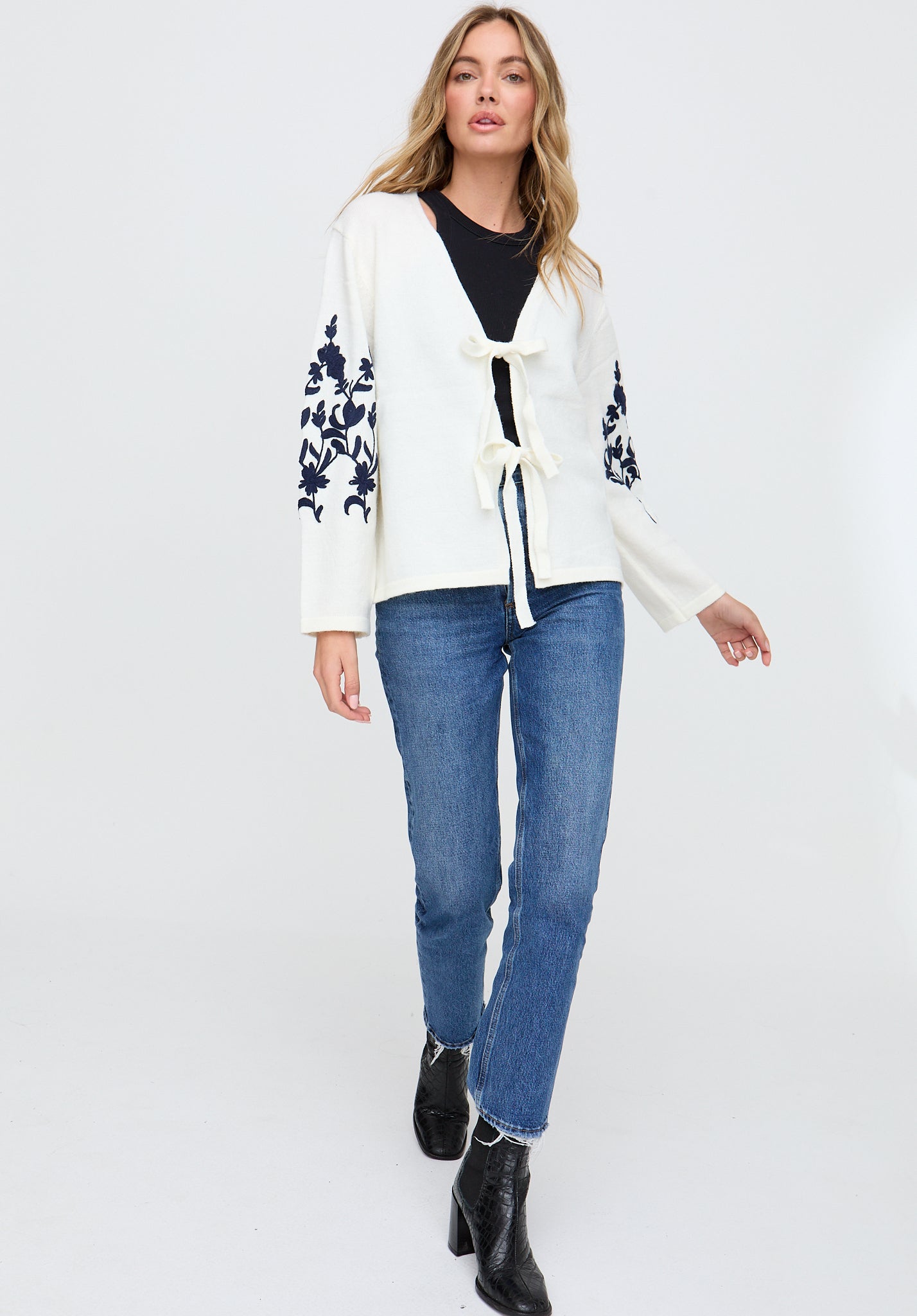Woman wearing a white cardigan with floral sleeves, black top, blue jeans, and black boots on a white background