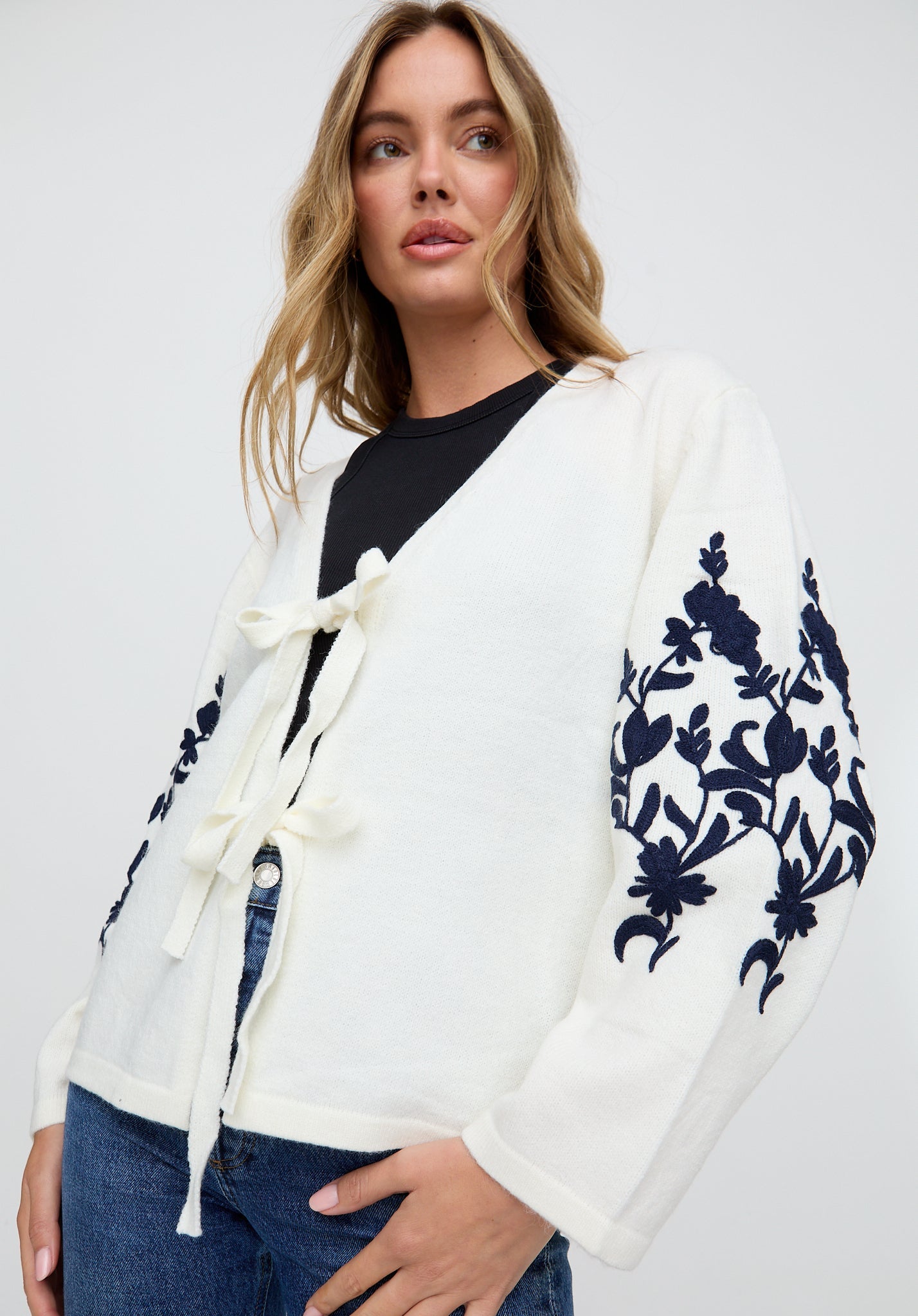 Woman wearing a white cardigan with blue floral embroidery on a plain background