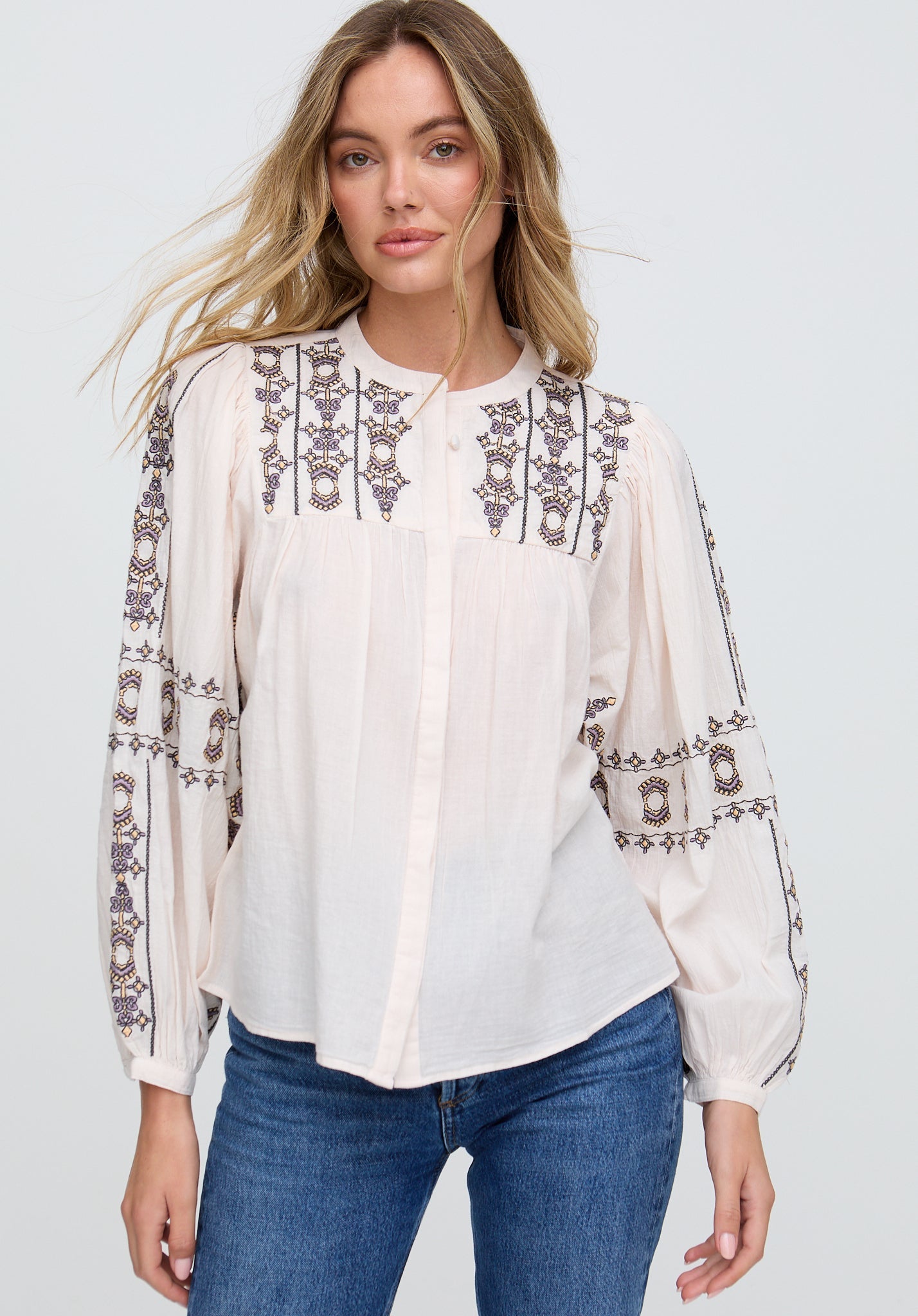 Woman wearing a white blouse with intricate black embroidery and blue jeans on a plain background